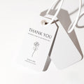 Minimal wedding favor tag featuring a refined serif thank you message, a delicate floral line illustration, and a soft natural shadow.