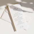 Editorial wedding menu featuring a long vertical layout with the handwritten title “Let’s eat,” printed on textured off-white paper and styled with layered envelopes and soft natural shadows.
