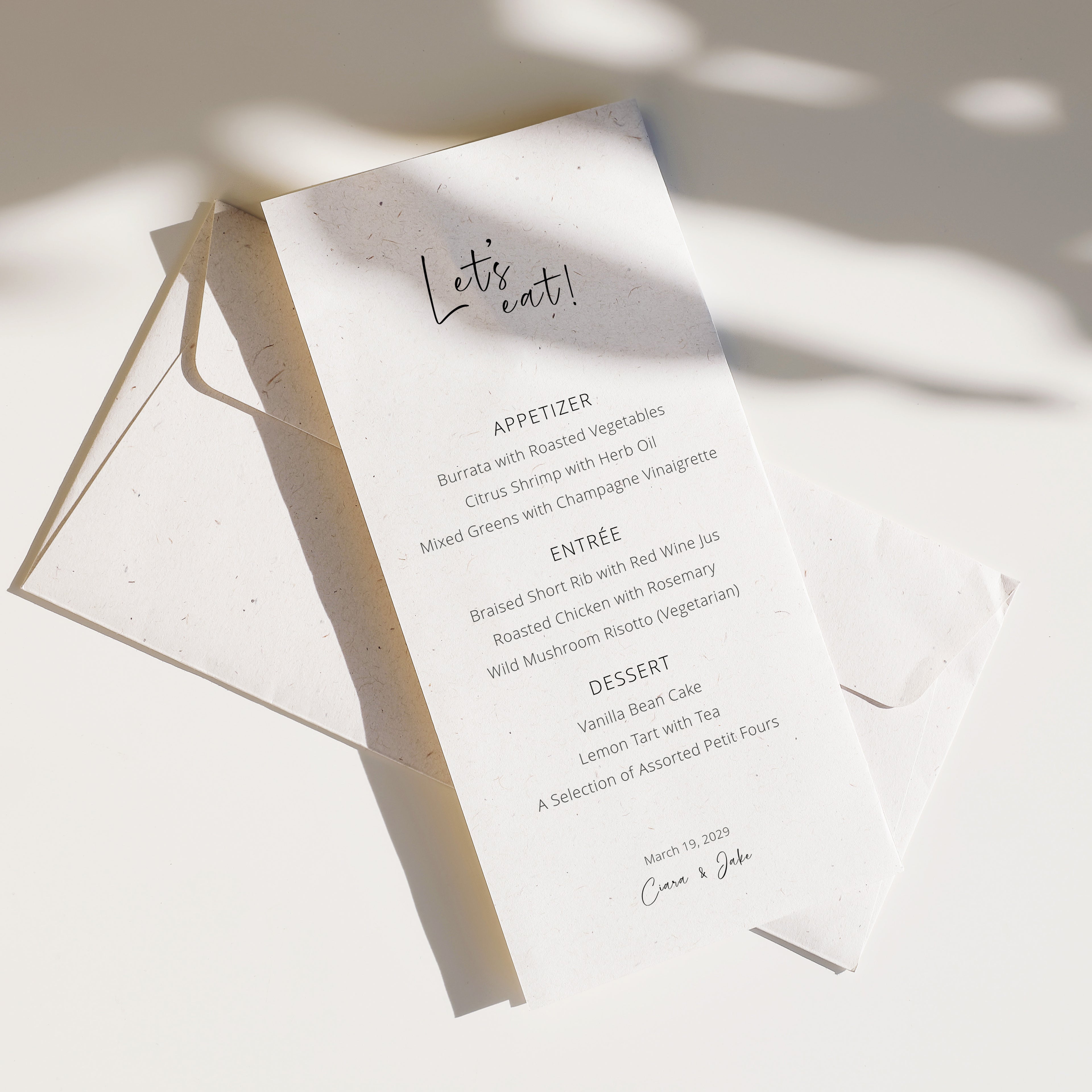 Editorial wedding menu featuring a long vertical layout with the handwritten title “Let’s eat,” printed on textured off-white paper and styled with layered envelopes and soft natural shadows.